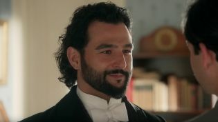 Mężczyzna w eleganckiej czarnej marynarce i białej koszuli z muszką uśmiecha się delikatnie, patrząc w dal. W tle widać rozmyte sylwetki innych postaci w klasycznym wnętrzu. Kadr z serialu „La Promesa – pałac tajemnic", gdzie arystokratyczne dramaty rozgr