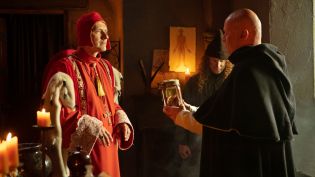 Kardynał Oleśnicki stoi w czerwonych szatach z futrem z gronostajów. Na głowie ma czerwoną czapkę. Inny duchowny podaje mu szklany słój. W tle świece i obraz człowieka. Odcinek 65 serialu Królowie dostępny jest w TVP VOD.