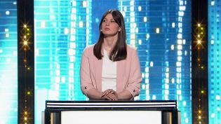 Zdjęcie z programu rozrywkowego „Jaka to melodia” dostępnego online w TVP VOD. Przedstawia uczestniczkę z długimi, brązowymi włosami, w białej koszuli i kremowym żakiecie. Trzyma dłonie na przycisku, a swój wzrok kieruje w lewą stronę. Tło stanowi fragmen