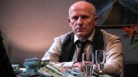 Mężczyzna w średnim wieku, ubrany w jasną koszulę, krawat i ciemny szalik z białymi kropkami, siedzi przy stole w serialu. Przed nim na stole leżą stosy banknotów euro, a obok stoją dwa kieliszki z wodą. W tle widoczne są kwiaty i rozmyte elementy wnętrza