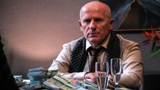 Mężczyzna w średnim wieku, ubrany w jasną koszulę, krawat i ciemny szalik z białymi kropkami, siedzi przy stole w serialu. Przed nim na stole leżą stosy banknotów euro, a obok stoją dwa kieliszki z wodą. W tle widoczne są kwiaty i rozmyte elementy wnętrza